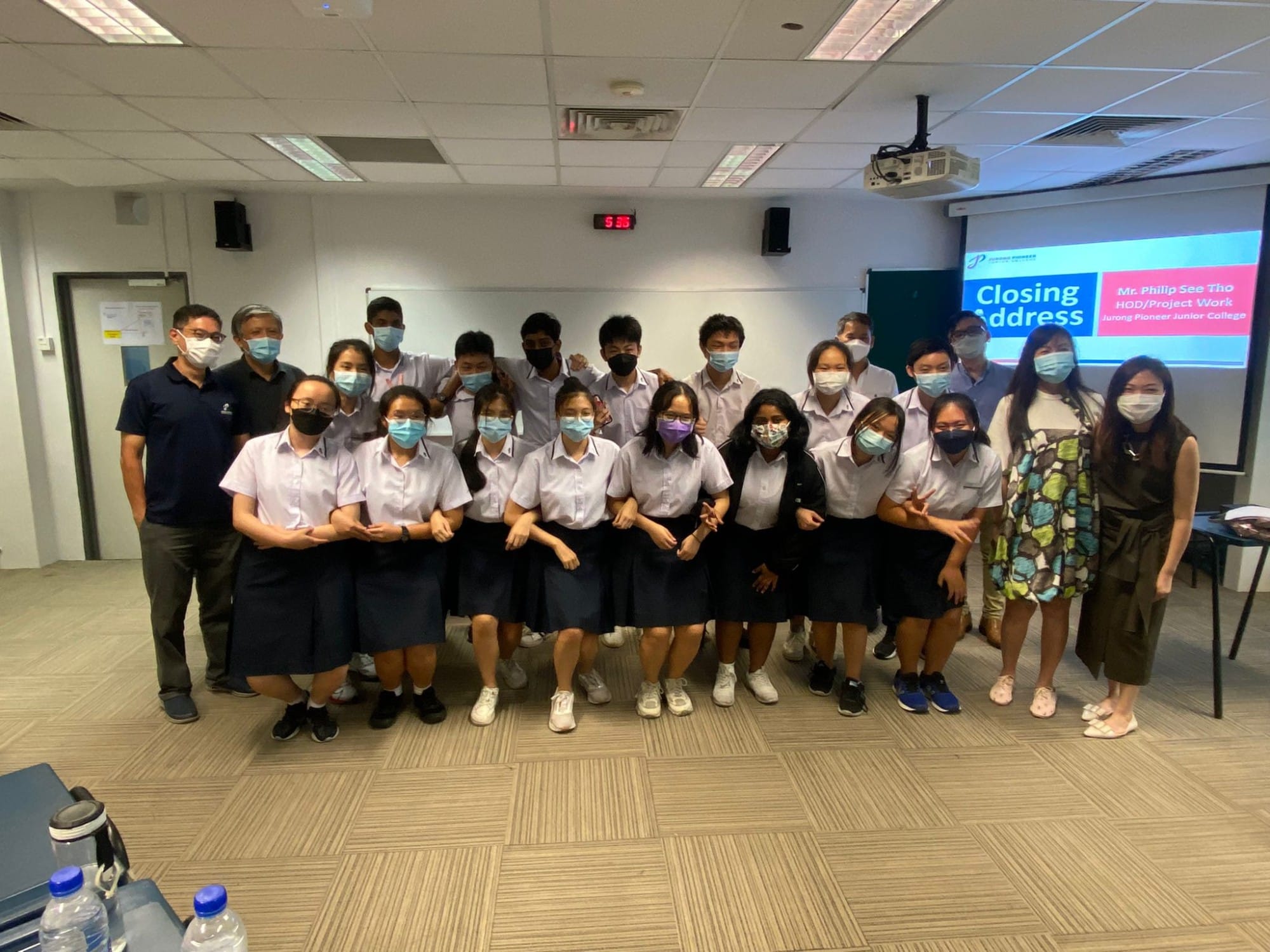 A group of masked students in school uniforms pose indoors near a projected "Closing Address" slide.